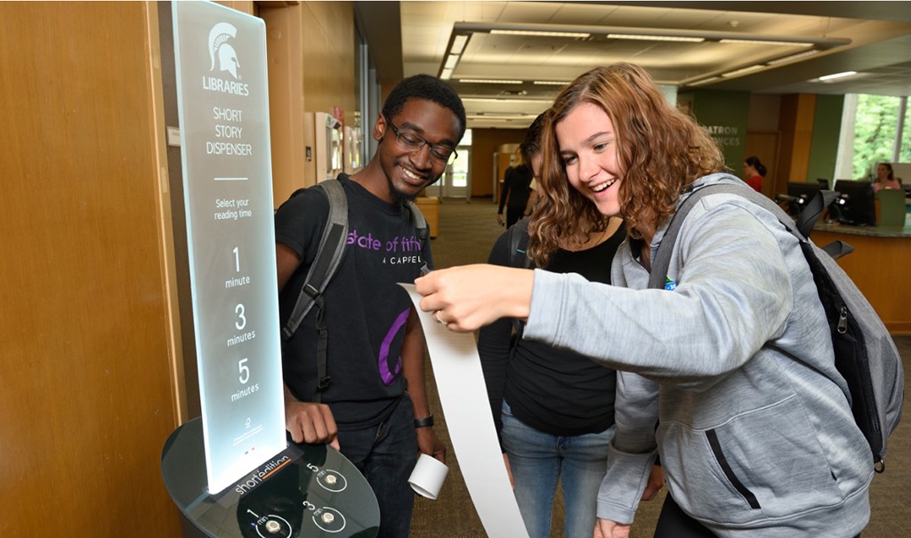 Students read a short story a dispenser printed on receipt paper in a library.