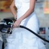 Woman from shoulders down wearing a white wedding dress shown refueling a car at a gas station left side view