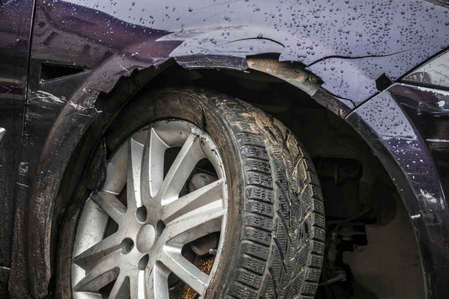 A car crash is shown in very close view dark colored car with damaged right front end is shown crooked wheel cracked fender