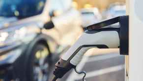 A white EV charger shown plugged into frame right while a blurred out car is parked with a charger plugged in the background