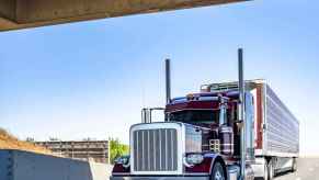 A classic red semi truck starts to pass under a bridge on the highway