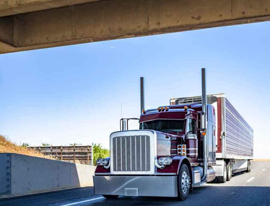 A classic red semi truck starts to pass under a bridge on the highway