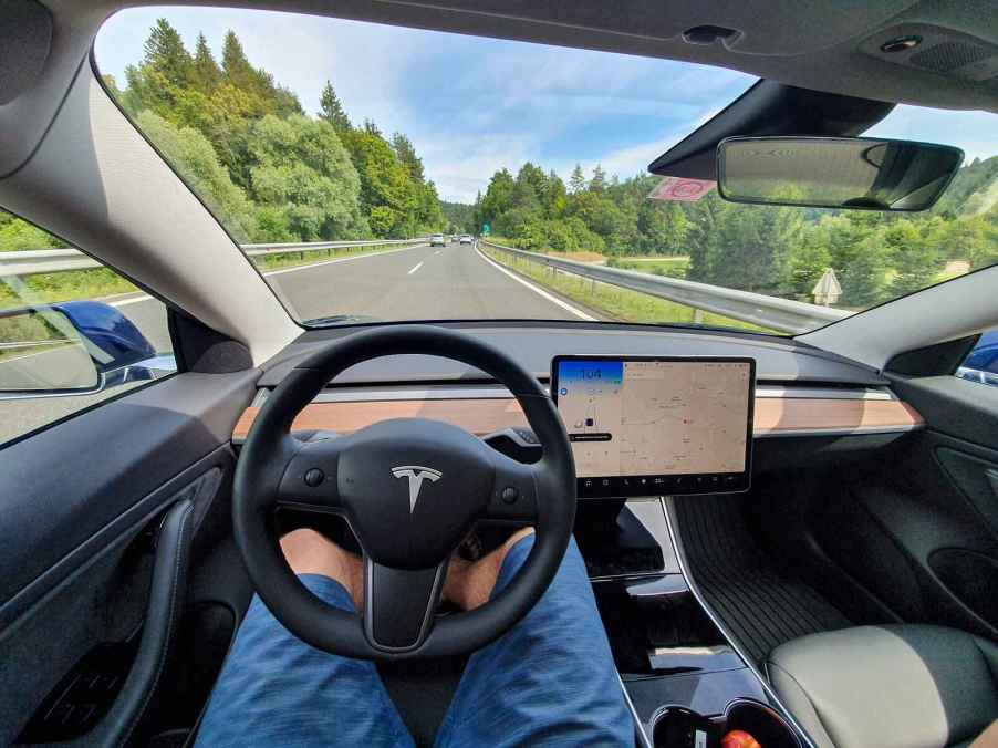 The driver interior view of a Tesla model in self driving mode man's legs under steering wheel no hands