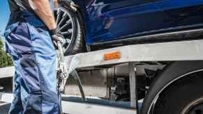 A tow truck driver dressed in blue work clothes is shown from neck to shins holding a tow strap next to a flatbed tow truck with a blue car on the bed