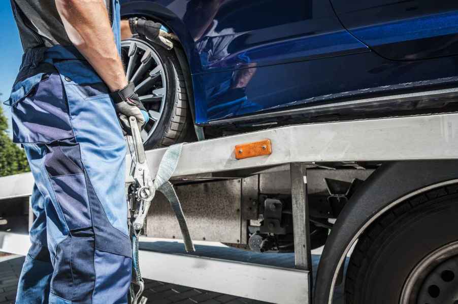 A tow truck driver dressed in blue work clothes is shown from neck to shins holding a tow strap next to a flatbed tow truck with a blue car on the bed