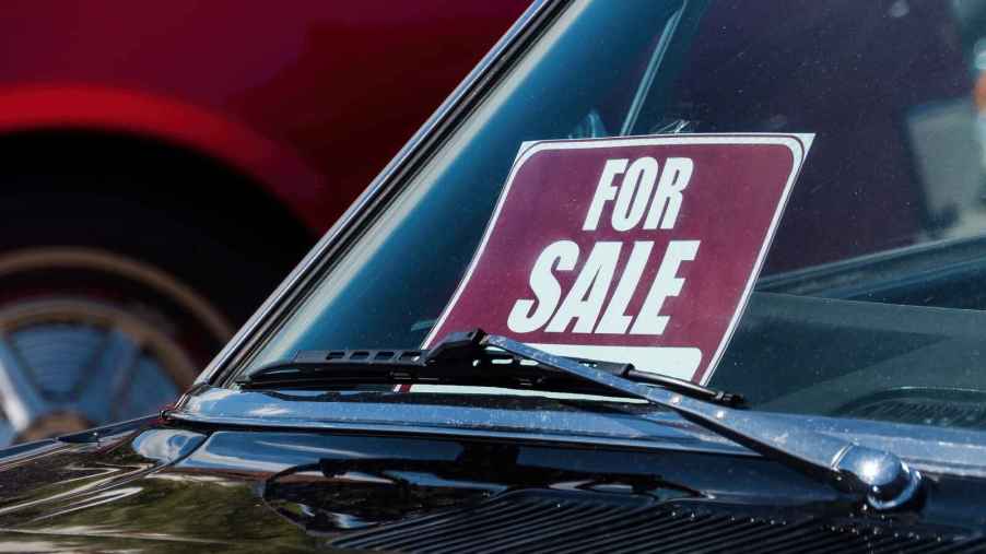 A red and white "FOR SALE" sign posted in a black car dash facing the windshield in close view.