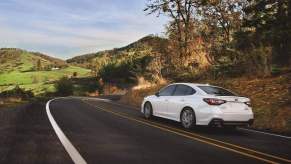 A white 2024 Subaru Legacy drives on a winding hilly road away from the viewer in left rear angle