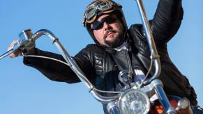 A rider rides with ape hangers on his custom motorcycle and what looks like a brain bucket helmet.