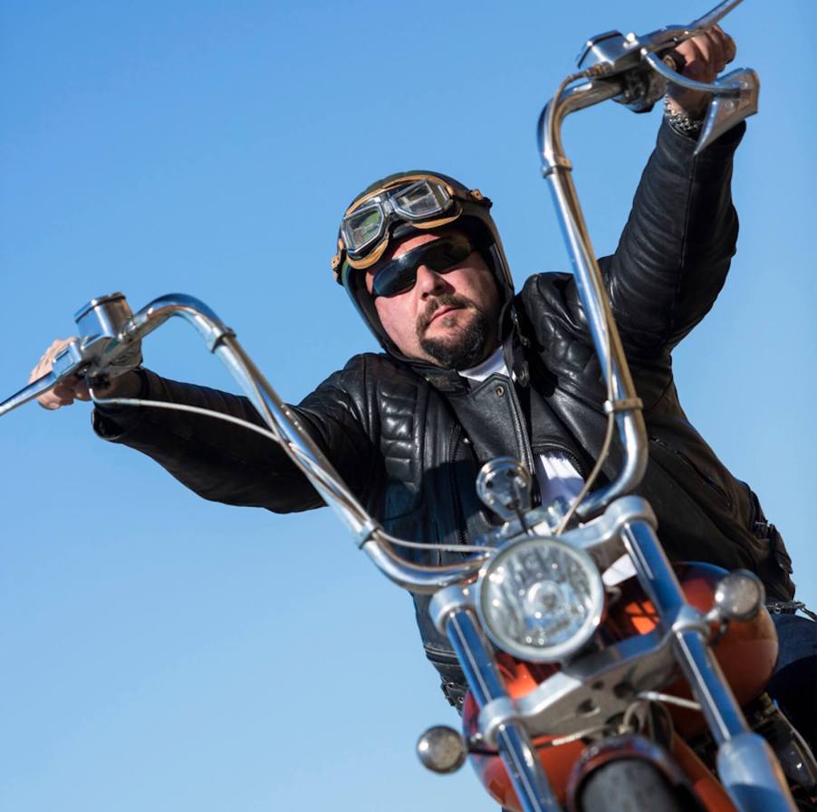 A rider rides with ape hangers on his custom motorcycle and what looks like a brain bucket helmet.