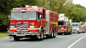 A parade of fire trucks cruise past a gas station on a busy street.