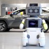 White robot with TV screen stands in front of a Hyundai EV at a dealership.