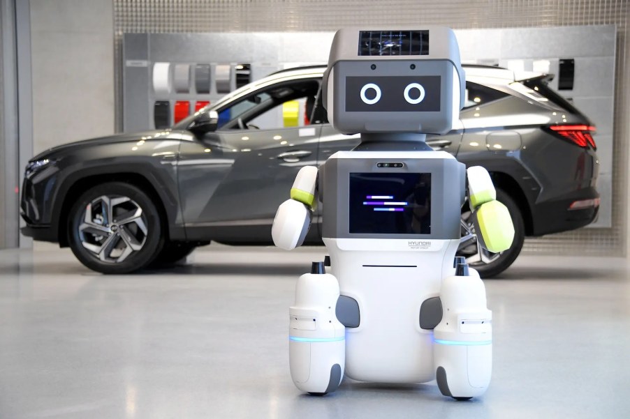 White robot with TV screen stands in front of a Hyundai EV at a dealership.