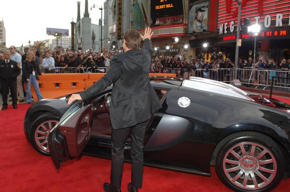 Tom Cruise stands next to his Bugatti Veyron on the red carpet and waves.