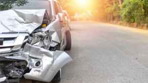 A silver car with a wrecked front end shown parked facing viewer left of center frame sun shining through trees background midframe