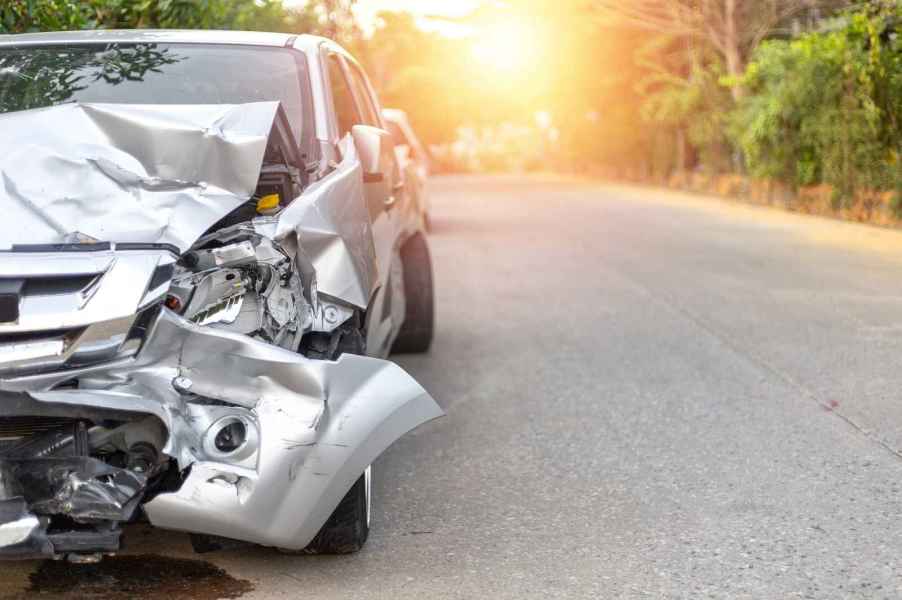 A silver car with a wrecked front end shown parked facing viewer left of center frame sun shining through trees background midframe