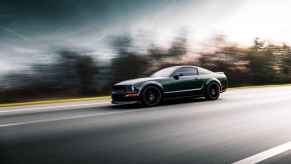 A dark colored Ford Mustang shown driving fast on a blurred road in left front profile view