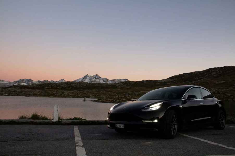 A black Tesla Model 3 parked in left front angle view with headlines glowing at deep dusk in front of water and snow capped mountain