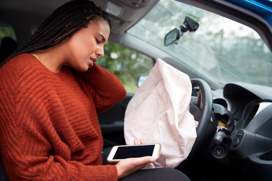 A woman in a crashed car with a deployed airbag rubs her neck and looks at her phone