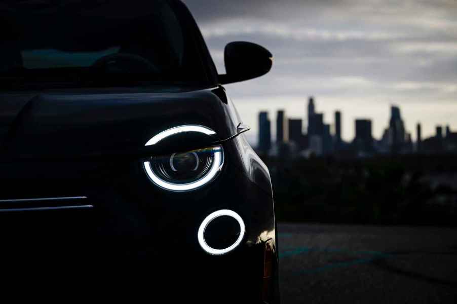 A 2024 Fiat 500e photographed outside in shadow with left headlamp illuminated in close left front side view with cityscape in right frame background