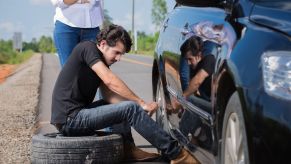 Man sits on his spare tire while changing a blown out tire on a summer roadtrip.