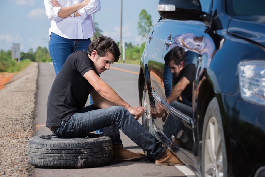 Man sits on his spare tire while changing a blown out tire on a summer roadtrip.