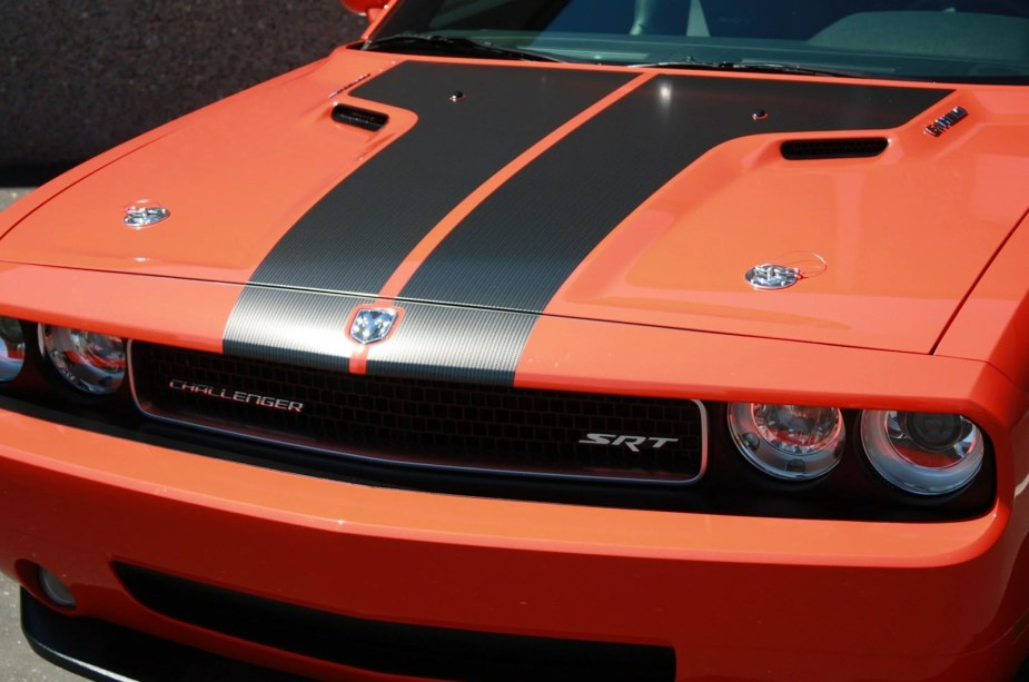 The hood of a bright orange Dodge Challenger with black racing strips and chrome hood pins.