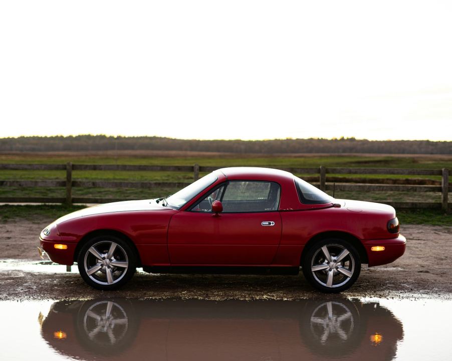 Red vintage Mazda Miata with an aftermarket hardtop coupe roof, parked in front of a green field.