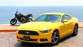 Bright yellow Mustang with mototcycle behind it and the ocean visible behind that.