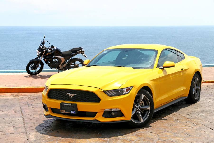 Bright yellow Mustang with mototcycle behind it and the ocean visible behind that.