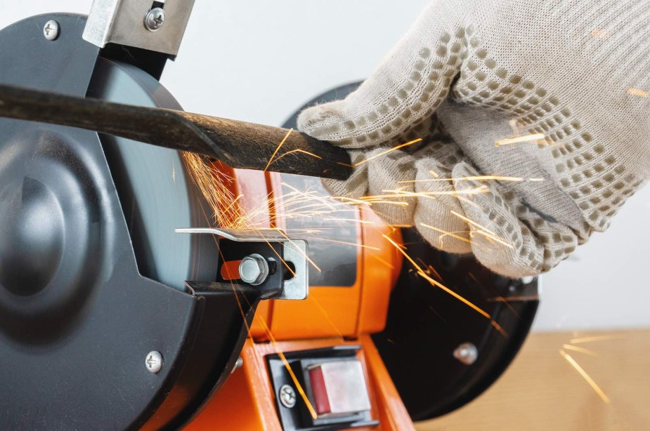 Gloved hands sharpening a mower blade on a bright orange grinder machine.