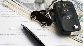 A key fob with car-shaped keychain sits on top of a stack of $100 bills near a black pen all on top of typed paper contract