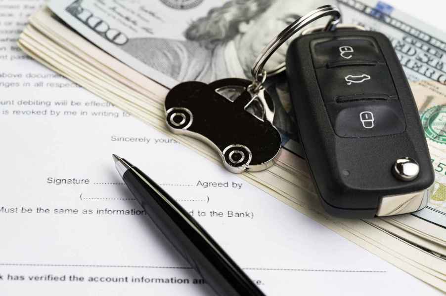 A key fob with car-shaped keychain sits on top of a stack of $100 bills near a black pen all on top of typed paper contract