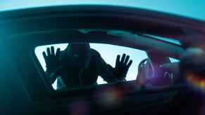 A man dressed in all black with black mask holds hands and face against left front car window clear view through front car windows