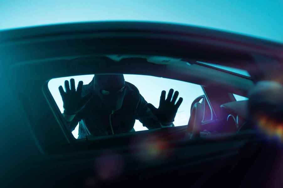 A man dressed in all black with black mask holds hands and face against left front car window clear view through front car windows