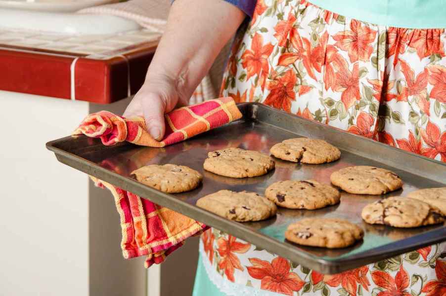 A 1950s-style close up of a woman holding a tray of chocolate chip cookies wearing a flowery apron