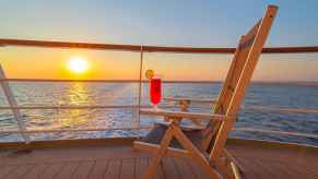 A wooden folding chair with pink cocktail on arm sits on a cruise ship deck at sunset