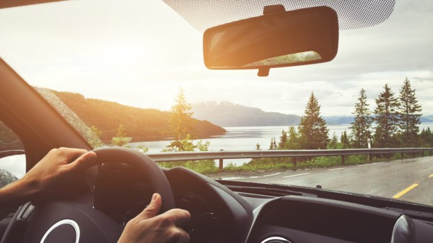 The view out of a black car interior driving with hands on steering wheel pine trees and lake in background paved road with guardrail in mid ground