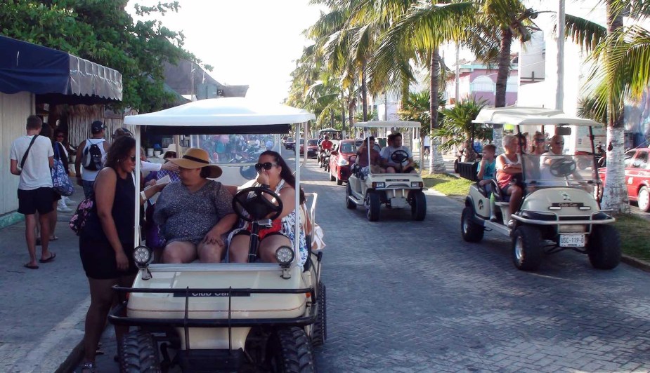 Golf cart drivers and passengers top on a public road to talk with a pedestrian, other golf carts and cars visible behind them.