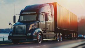 A semi-truck in close left front angle view driving on a highway with blurry sun in right upper corner