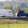 A black SUV with roof gear box towing a light colored camper with evergreen trees in the background