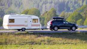 A black SUV with roof gear box towing a light colored camper with evergreen trees in the background