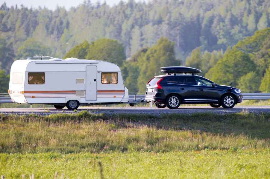 A black SUV with roof gear box towing a light colored camper with evergreen trees in the background