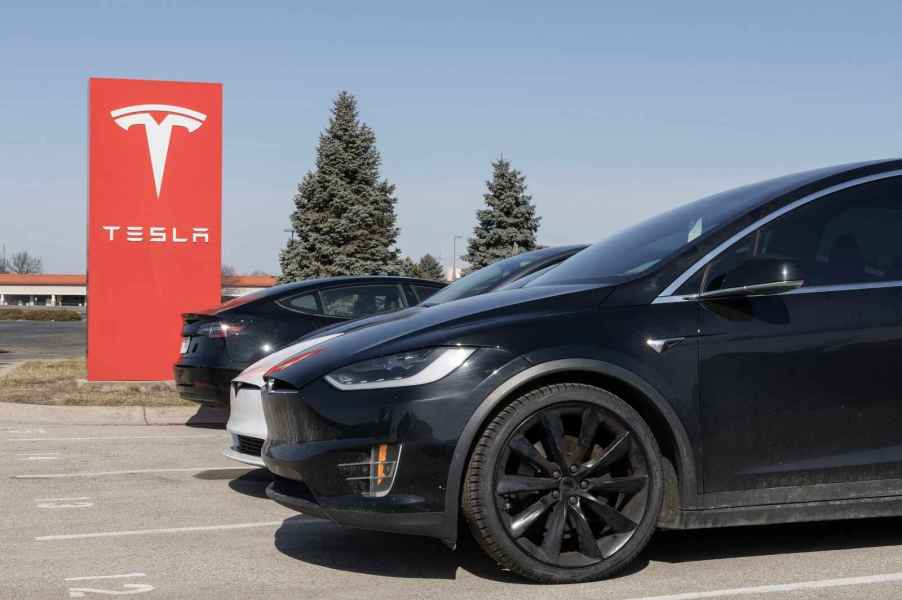 A red Tesla sign with a row of Tesla cars parked in a lot