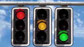 Three traffic light signals each showing one color, red, yellow, green, hang in a row with cloudy blue sky in the background