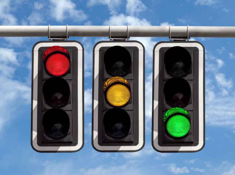 Three traffic light signals each showing one color, red, yellow, green, hang in a row with cloudy blue sky in the background