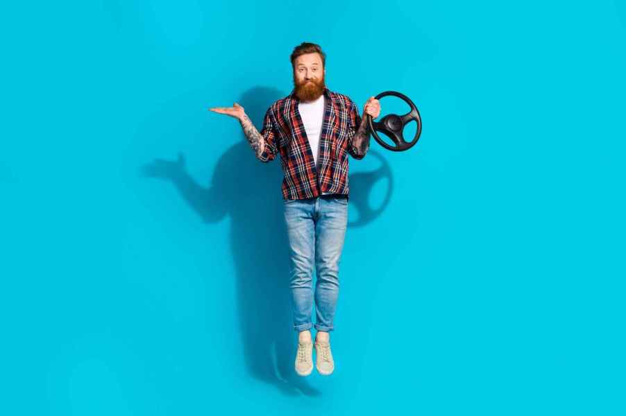 A man with a long beard dark red hair jumping in front of a blue screen holding a steering wheel prop in one hand with other arm in a palm-up shrug position