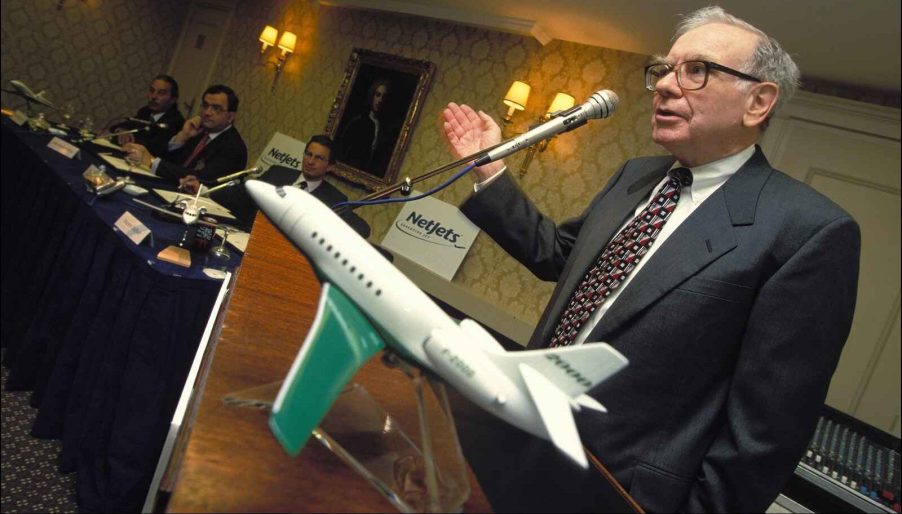 Warren Buffett speaking at a podium with NetJets signage in background and NetJets model airplane next to him