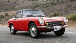 A red 1965 Honda S600 convertible roadster on a road with a grassy hill in the background