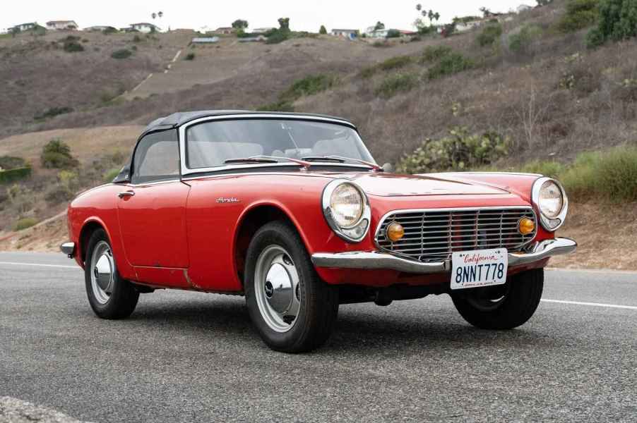 A red 1965 Honda S600 convertible roadster on a road with a grassy hill in the background