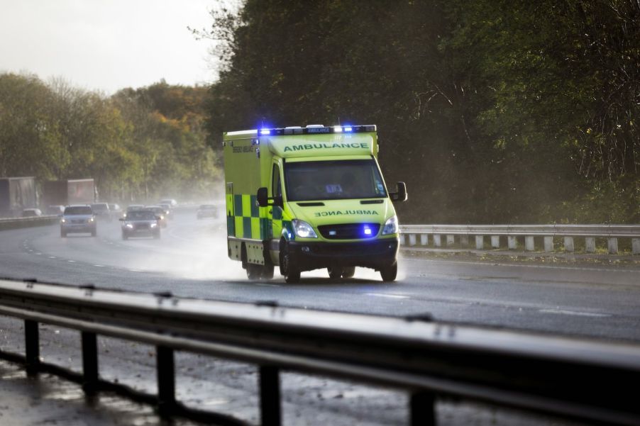 A neon yellow checkered British ambulance races down a UK highway in the rain.